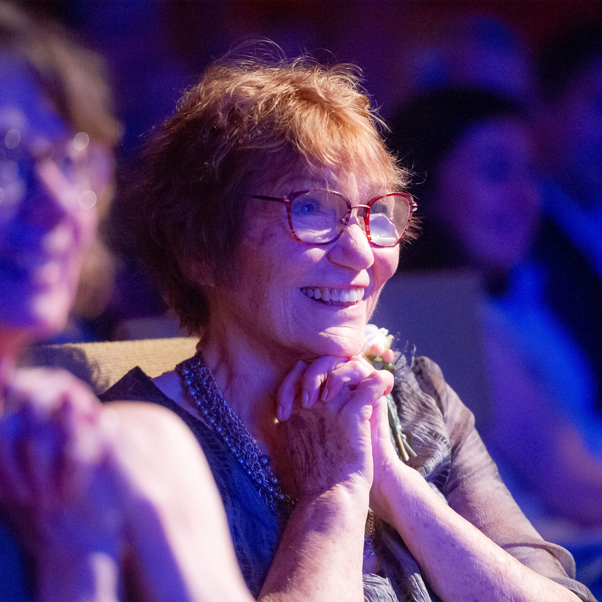 a woman with short red hair and glasses holds her hands to her heart and smiles. She is seated in a theater.