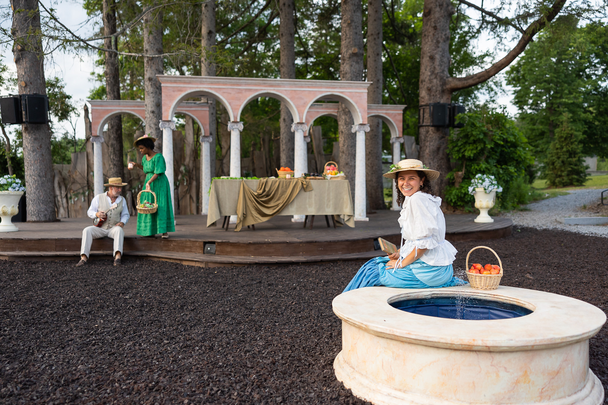 A woman sits outside on a fountain smiling, with two people - a man in a vest and a woman in a green dress - behind her.